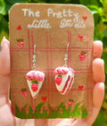 Pair of strawberry-themed earrings on a decorative card held by a hand with a natural background.