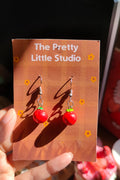 Red berry earrings on a 'The Pretty Little Studio' card held by a hand.